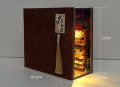 Miniature book nook kit with dark fabric-textured cover and tassel, open side showing lit interior