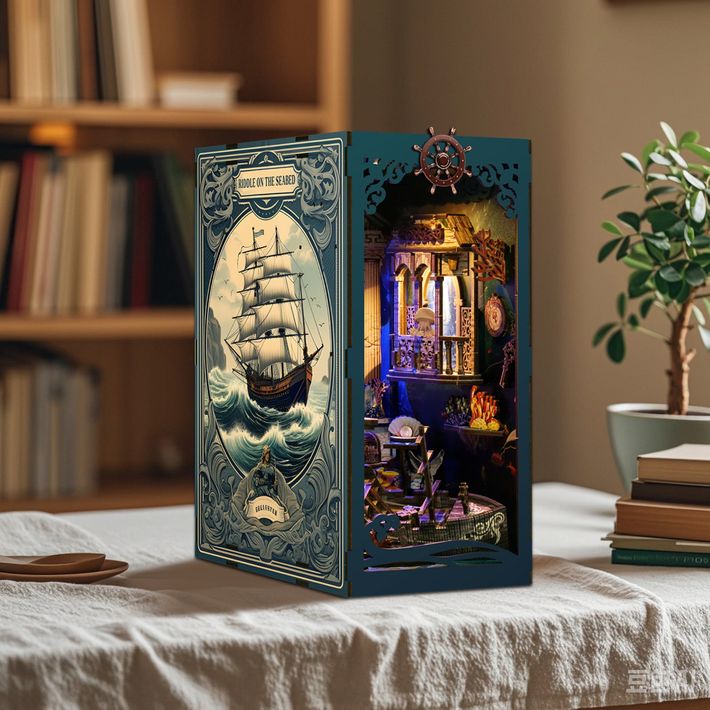 a miniature ship model with a lighthouse and a small ship, displayed on a table with books and a plant in the background.