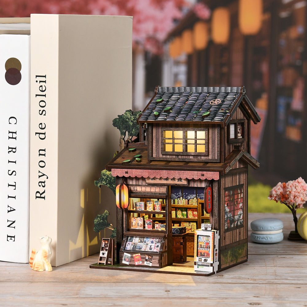 Miniature wooden grocery store model next to kit box, with lit shopfront windows