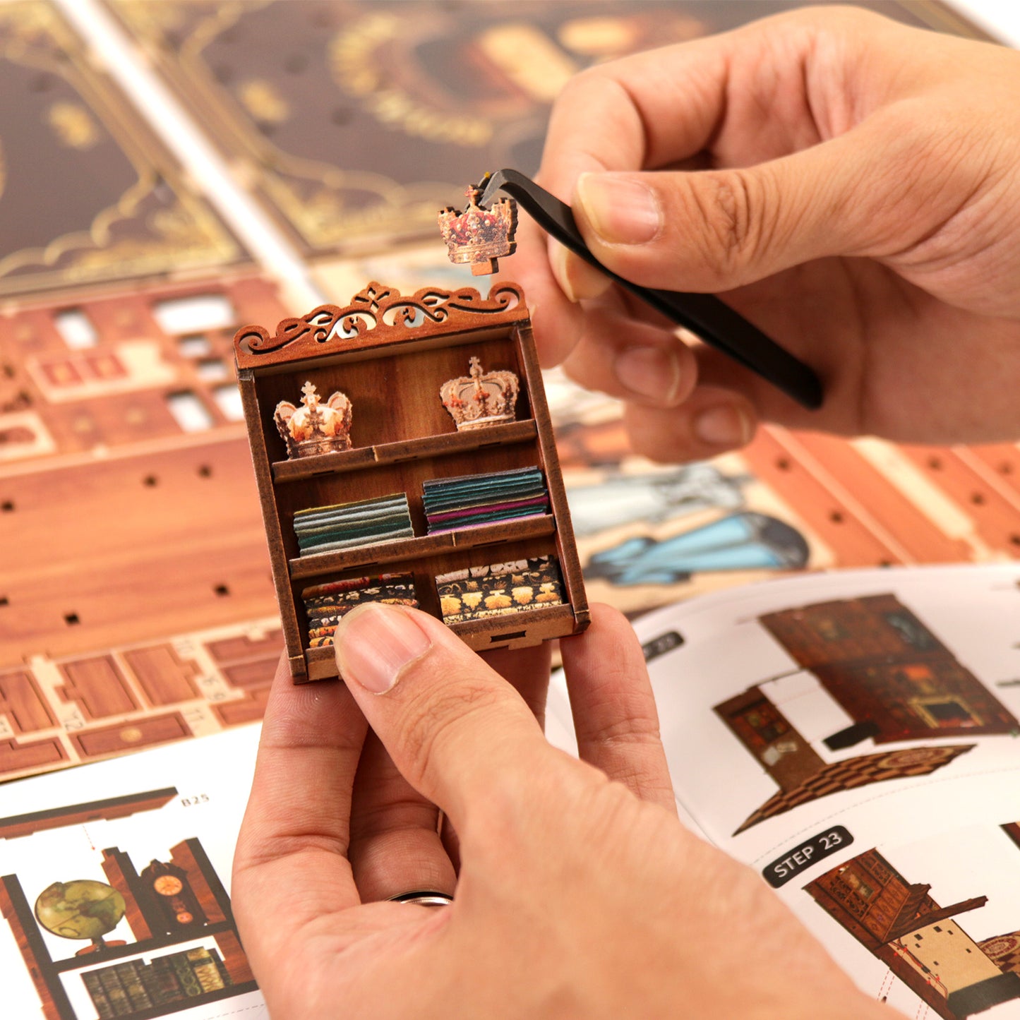 A person's hands are carefully assembling a miniature wooden bookshelf, with a small crown on top of the shelf.