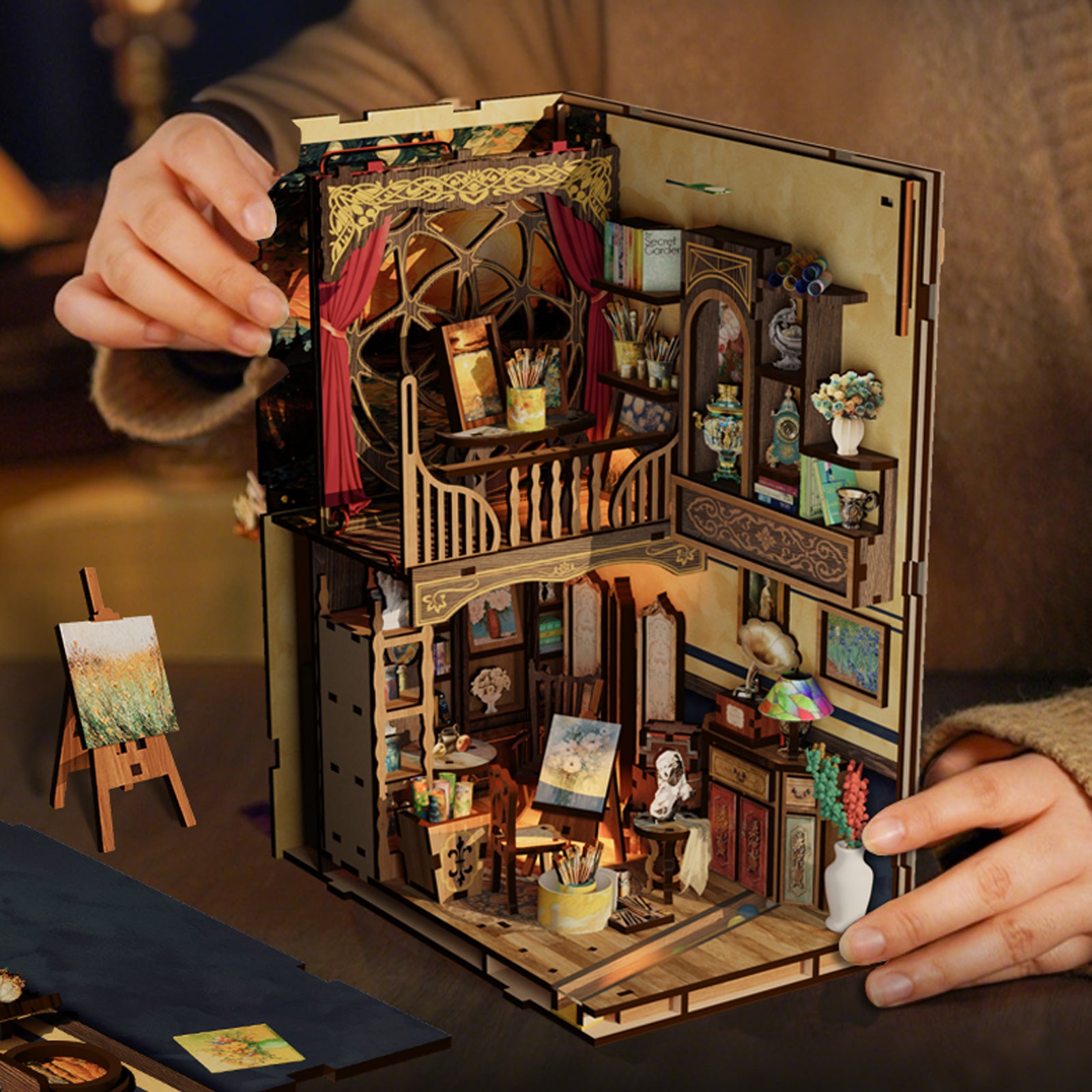 A person is holding a miniature wooden dollhouse, which appears to be a detailed and intricate model of a home or studio.
