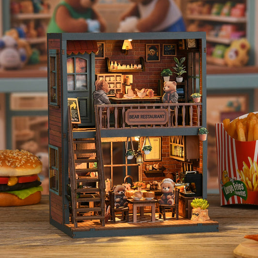 A miniature model of a bear-themed restaurant, complete with a dining area, kitchen, and seating for several teddy bears. The restaurant is decorated with various elements such as potted plants, a sign, and a menu board. The scene is set on a table, and there are also some food items, including a hamburger and a container of fries, placed nearby.