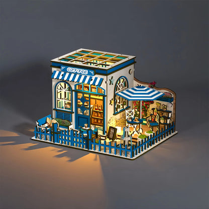A miniature model of a coffee shop, featuring a blue and white exterior, a small patio with a blue umbrella, and various decorative elements such as potted plants and a sign.