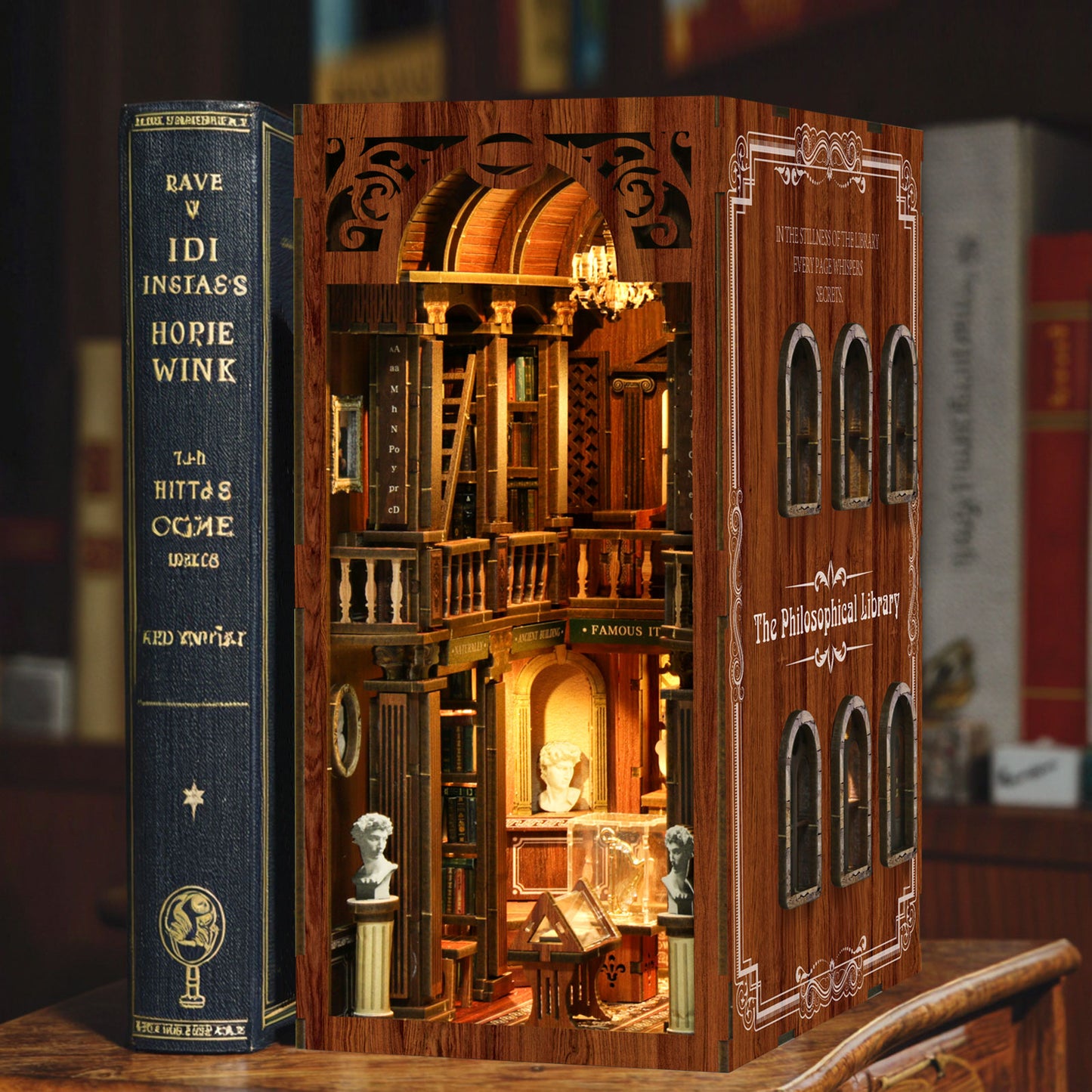 a miniature dollhouse-like structure with a detailed interior, resembling a book. The structure is made of wood and has intricate architectural elements, such as arches and columns. The dollhouse appears to be a miniature replica of a library or bookstore, with bookshelves and a chandelier. The structure is placed on a wooden surface, and there are books visible in the background, suggesting the scene is set in a library or bookstore.