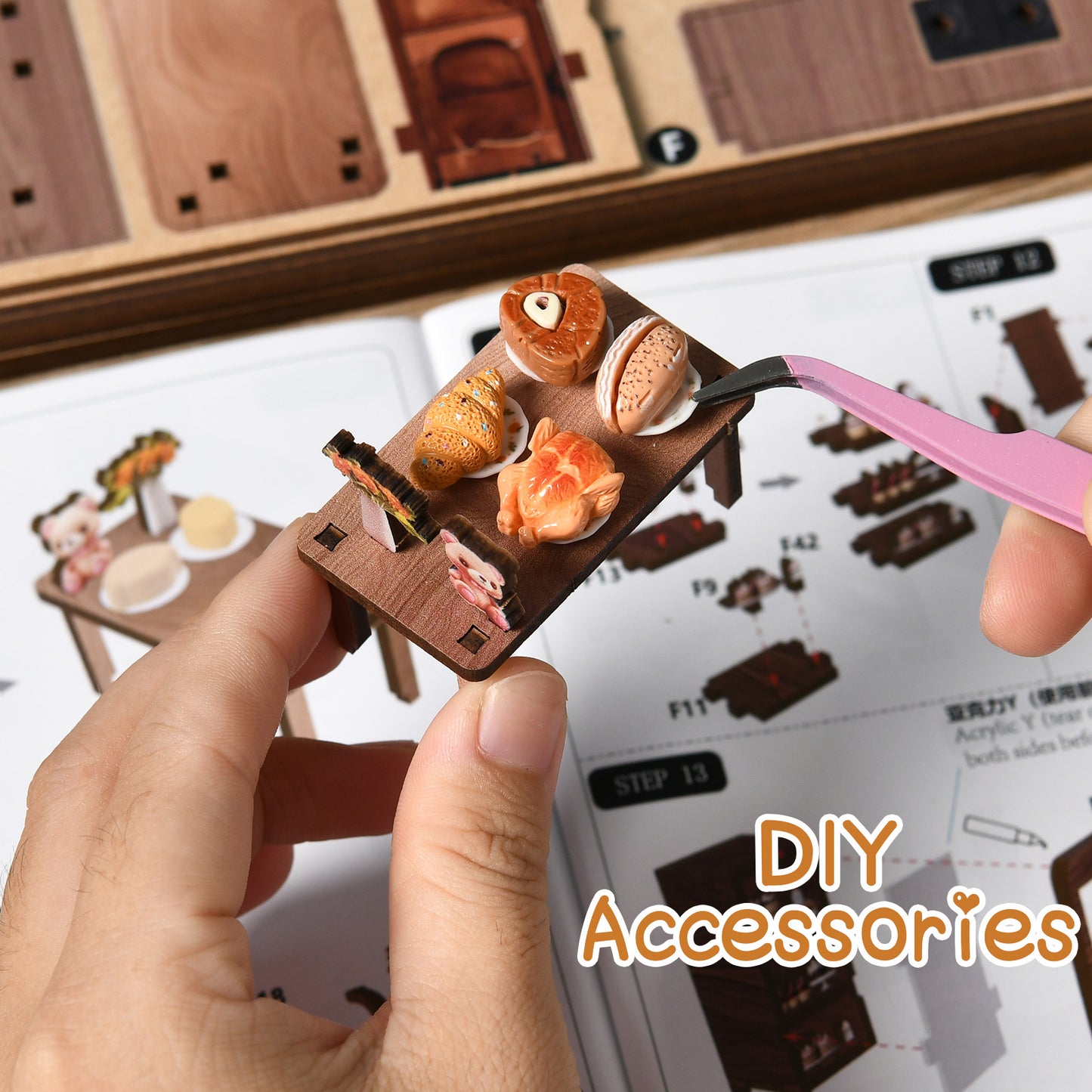 A person's hand holding a small wooden tray with various food items, such as a croissant, bagel, and sandwich, along with a pink tweezer. The hand is also holding a book or instructions for assembling a DIY accessory.