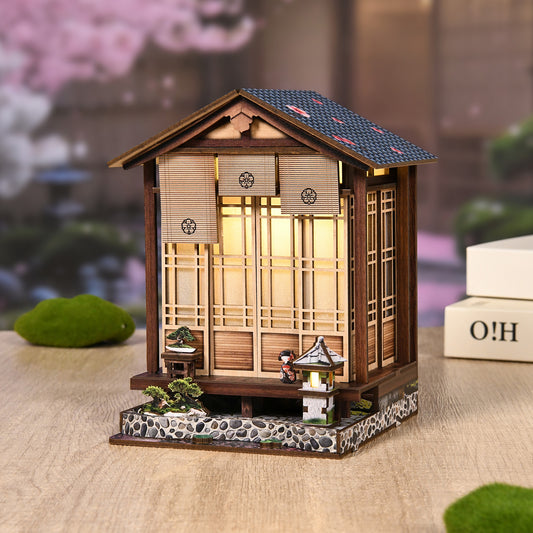A miniature wooden Japanese-style house with a tiled roof, sitting on a small stone base, surrounded by a garden scene with a small tree and a small lantern.