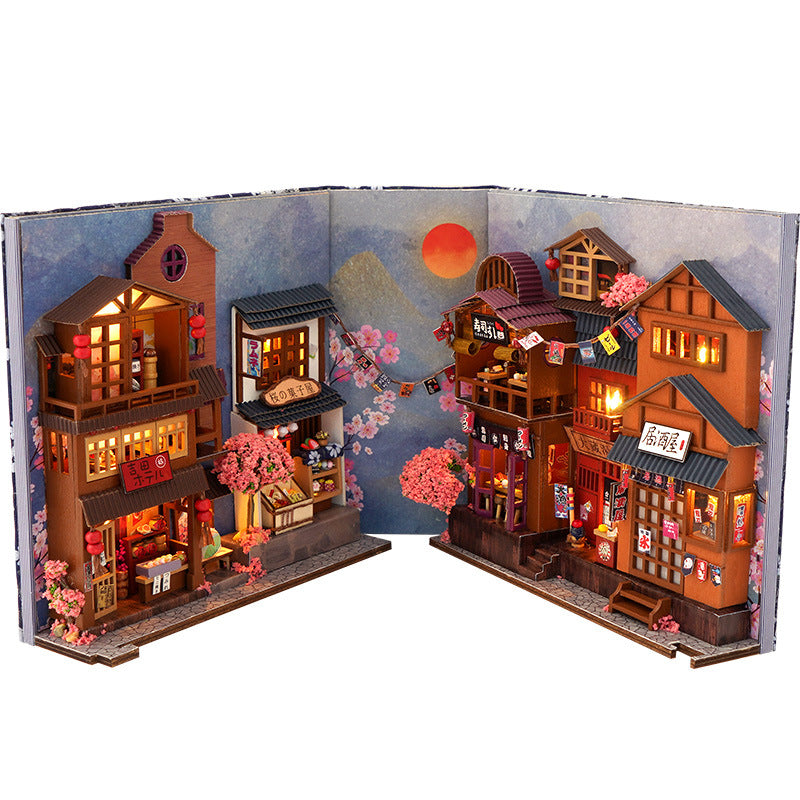Folded open Japanese-style book nook diorama with warm-lit storefronts and trees
