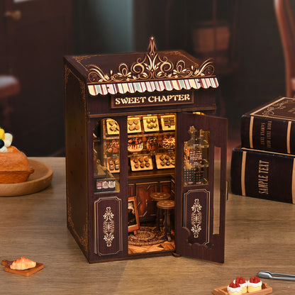 A miniature model of a small bakery shop, with a sign that reads "Sweet Chapter", displayed on a wooden table.