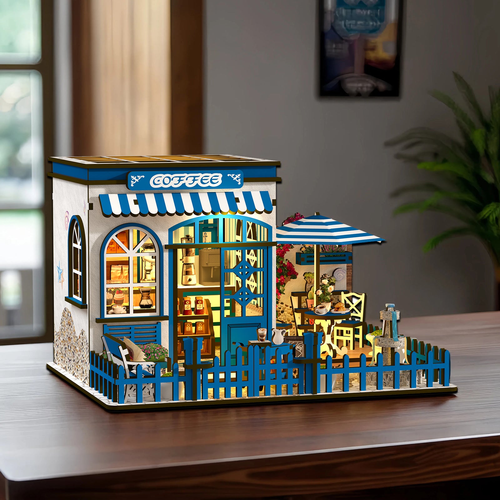 A miniature model of a cozy coffee shop, complete with a small outdoor seating area, a blue and white striped awning, and a charming wooden fence.