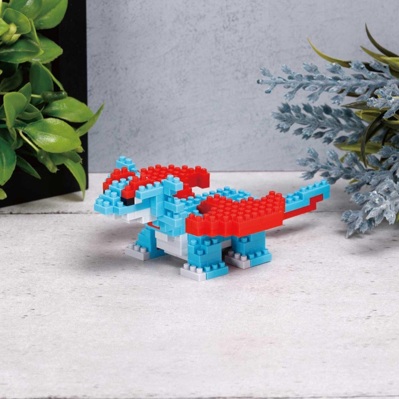 A small, colorful LEGO dragon figurine is sitting on a white surface, surrounded by greenery.