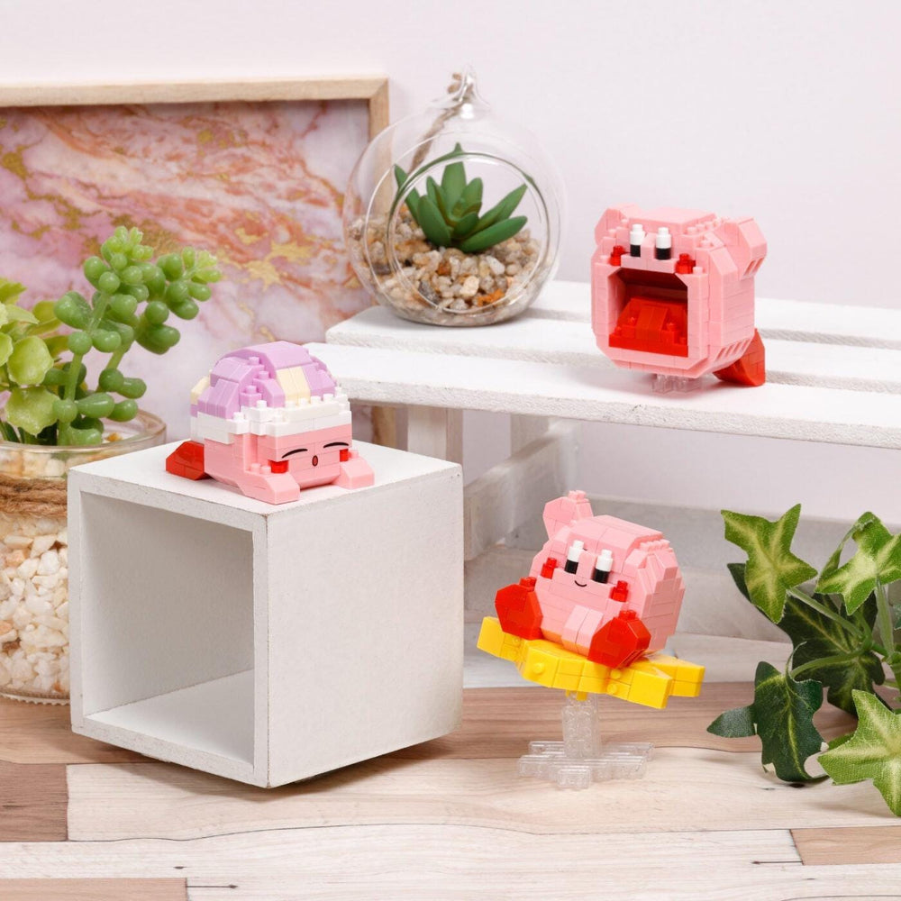 a collection of three small, colorful, and whimsical toys, including a pink and purple character, a pink and yellow character, and a pink and yellow character, all sitting on a white surface. The toys are surrounded by various decorative elements, such as a potted plant, a glass vase, and a framed picture, creating a playful and visually appealing scene.