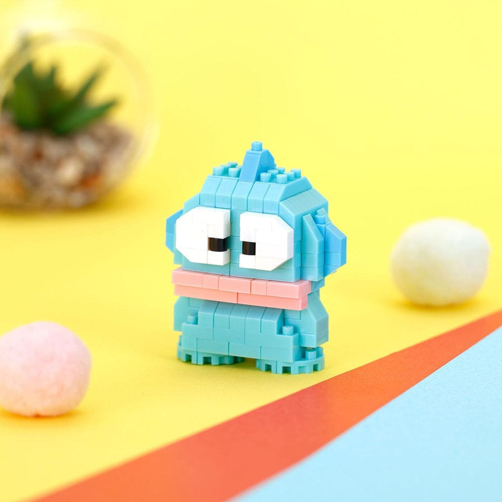 A small, colorful, and whimsical toy robot made of small building blocks is sitting on a yellow background, with a glass container of greenery and two small balls nearby.
