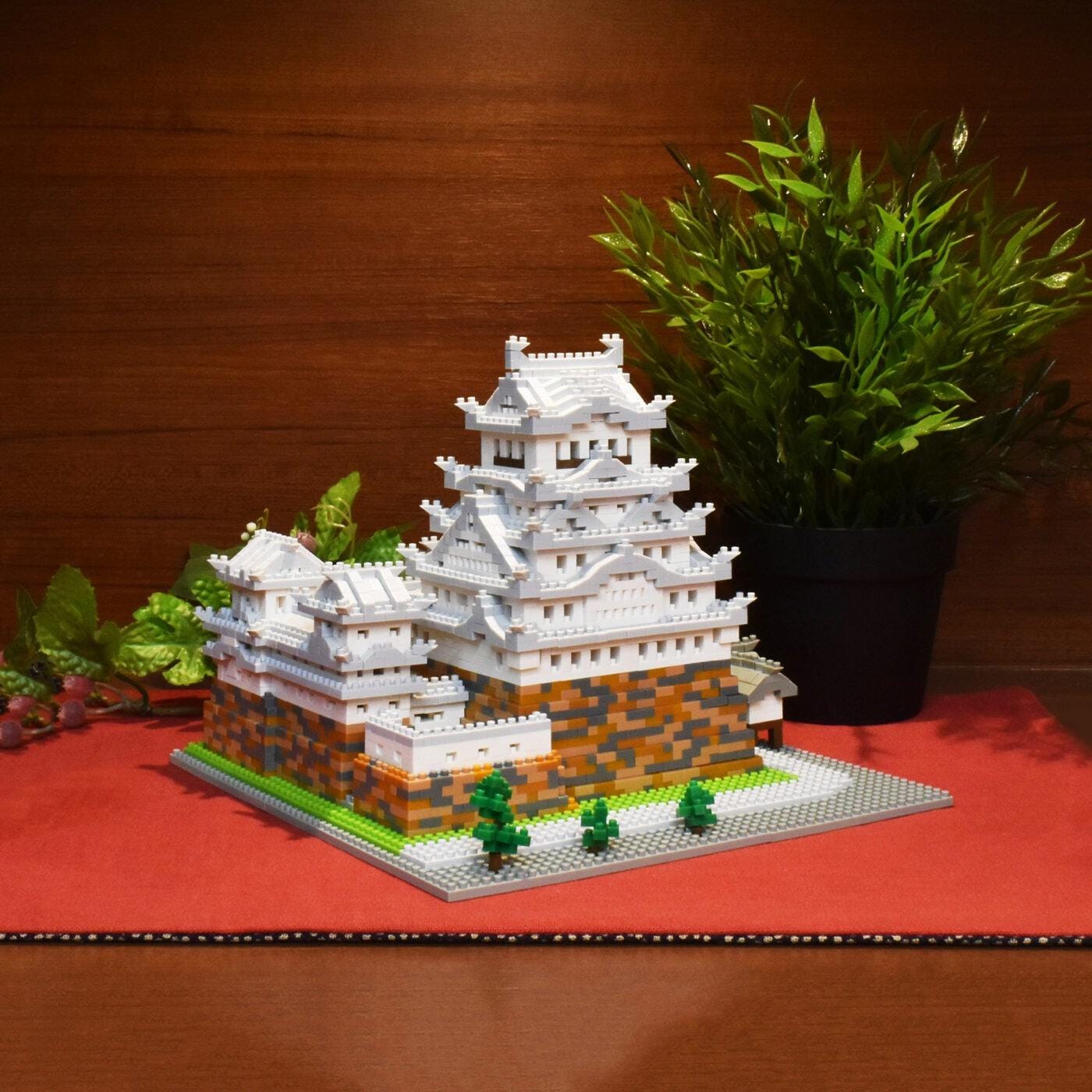 A miniature model of a Japanese castle made of white and orange building blocks, placed on a red surface next to a potted plant.