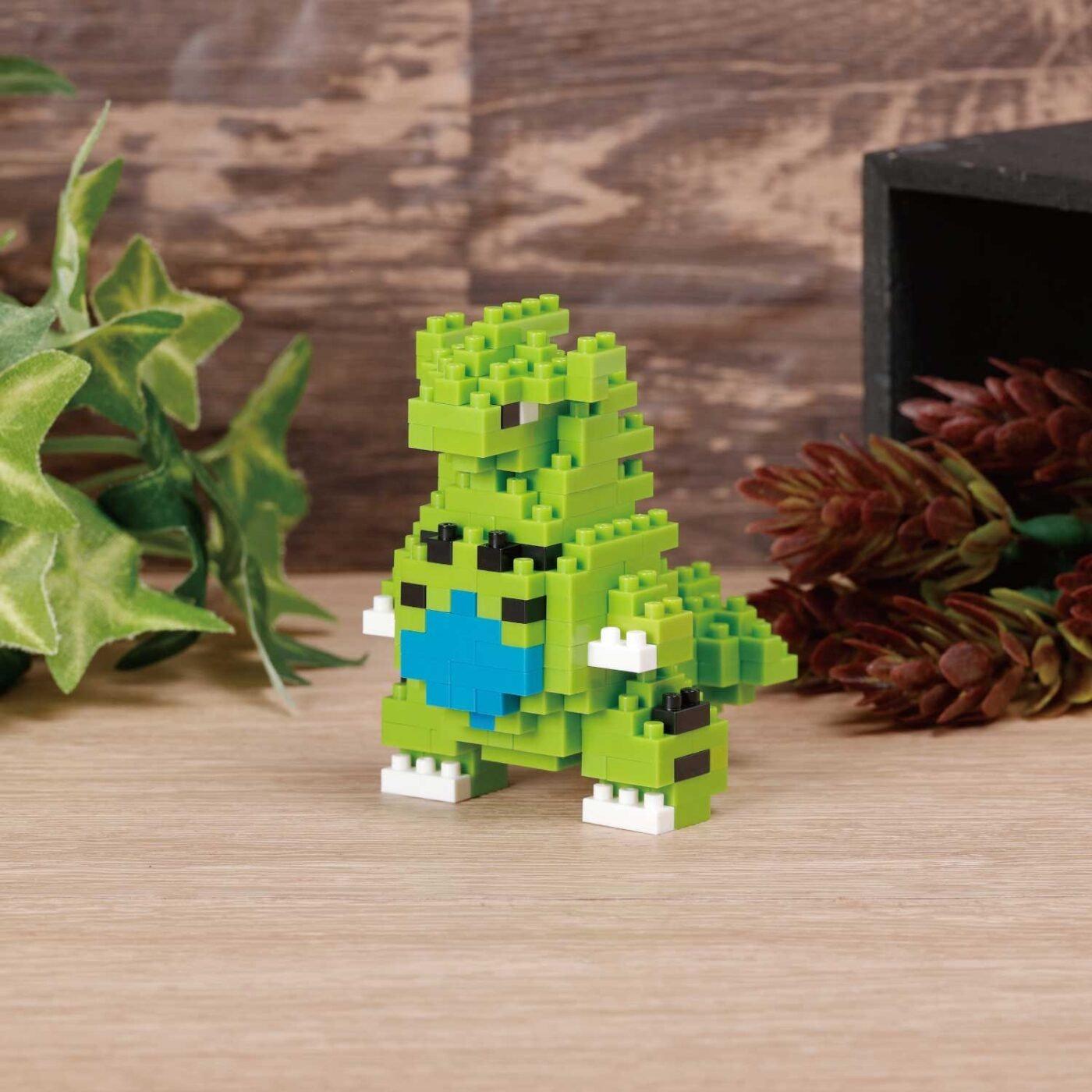 A small green LEGO dinosaur figurine is sitting on a wooden surface, surrounded by green leaves and pine cones.