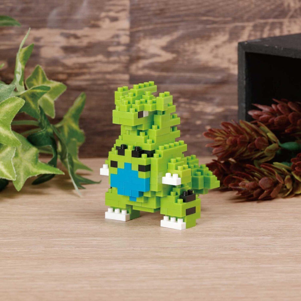 A small green LEGO dinosaur figurine is sitting on a wooden surface, surrounded by green leaves and pine cones.