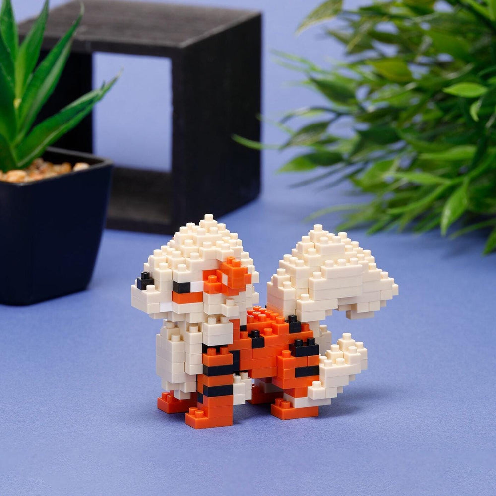 A small, colorful, and detailed toy resembling a pixelated character is displayed on a blue surface, with a potted plant and a black box in the background.