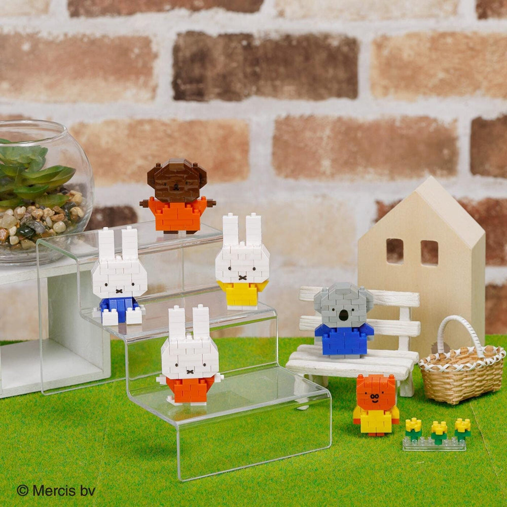A collection of colorful, small toy animals, including a bear, rabbit, and koala, arranged on a green surface. There is also a small house and a glass container with plants in the background.