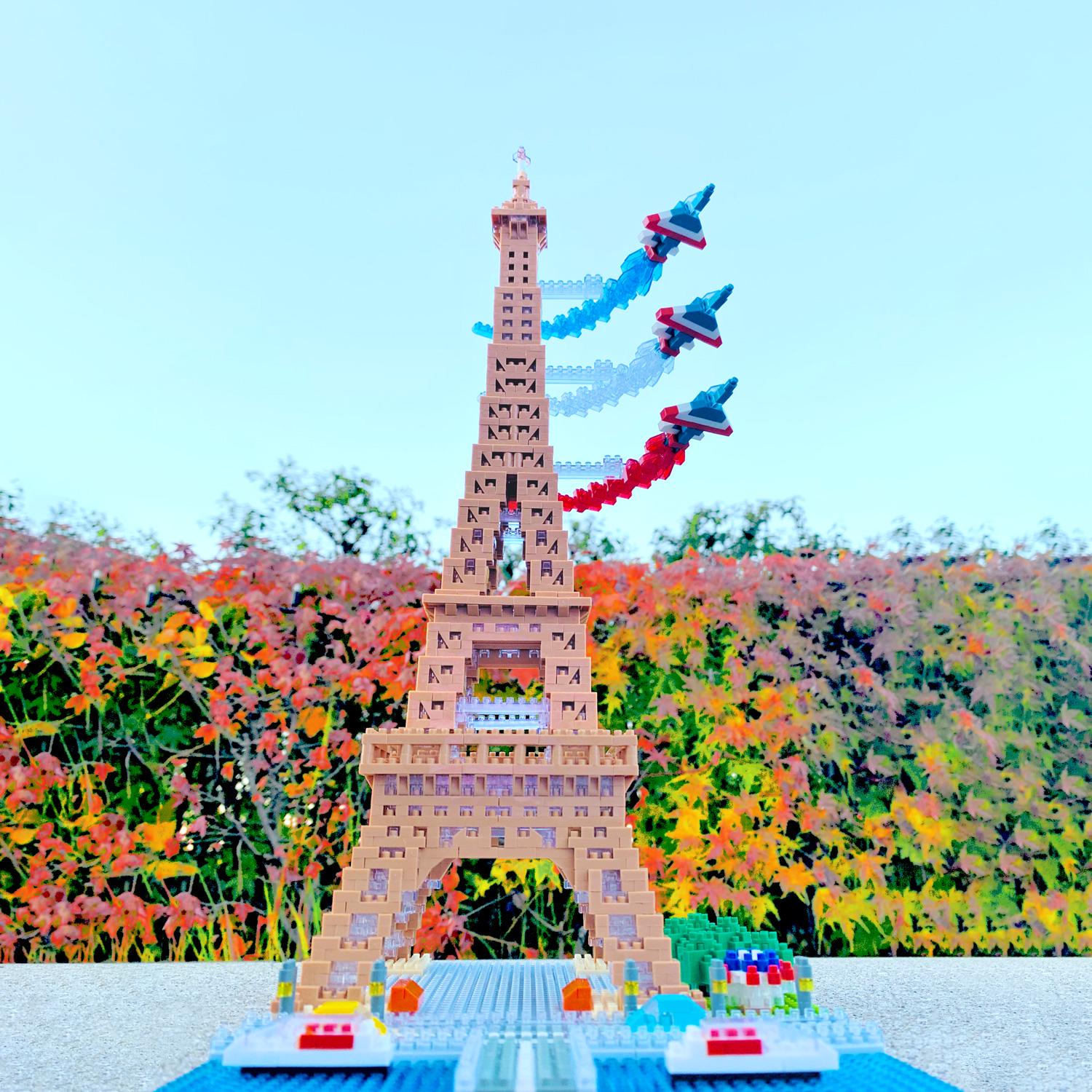 A colorful Lego tower with three Lego planes flying above it, set against a backdrop of a garden with autumn foliage.