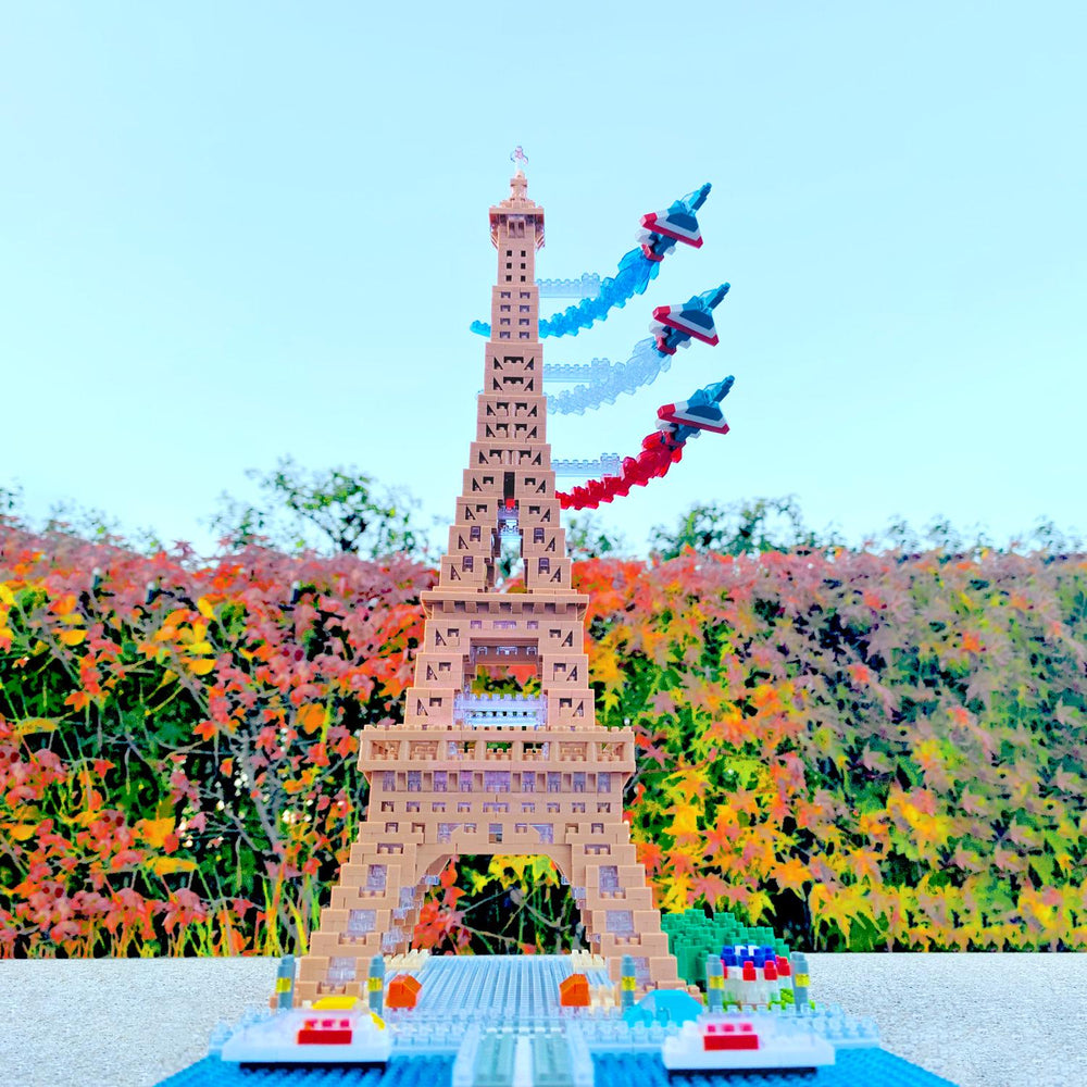 A colorful Lego tower with three Lego planes flying above it, set against a backdrop of a garden with autumn foliage.