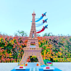 A colorful Lego tower with three Lego planes flying above it, set against a backdrop of a garden with autumn foliage.