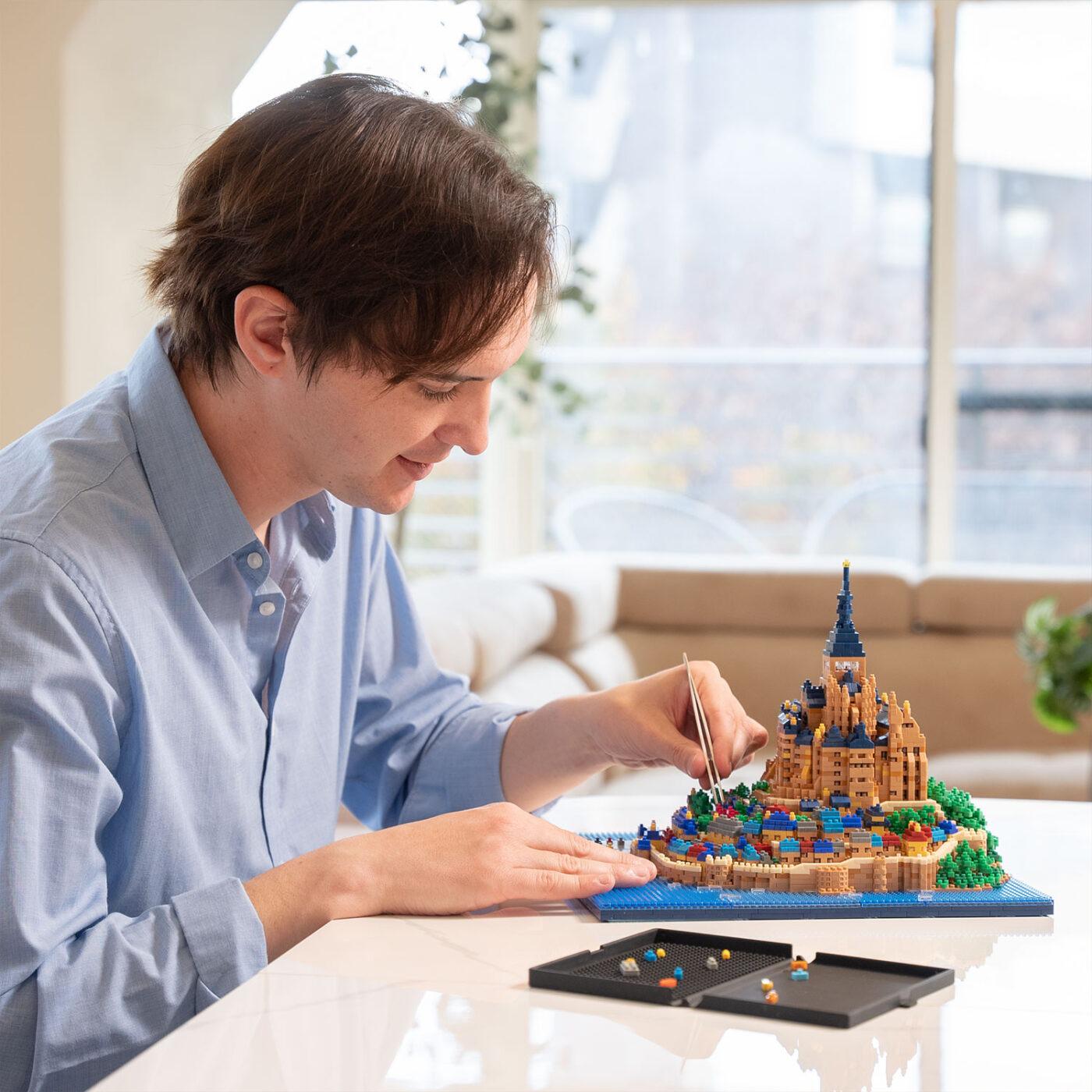 A man is sitting at a table, working on a small Lego castle model. He is carefully placing small Lego bricks onto the castle, which is a detailed and intricate model.