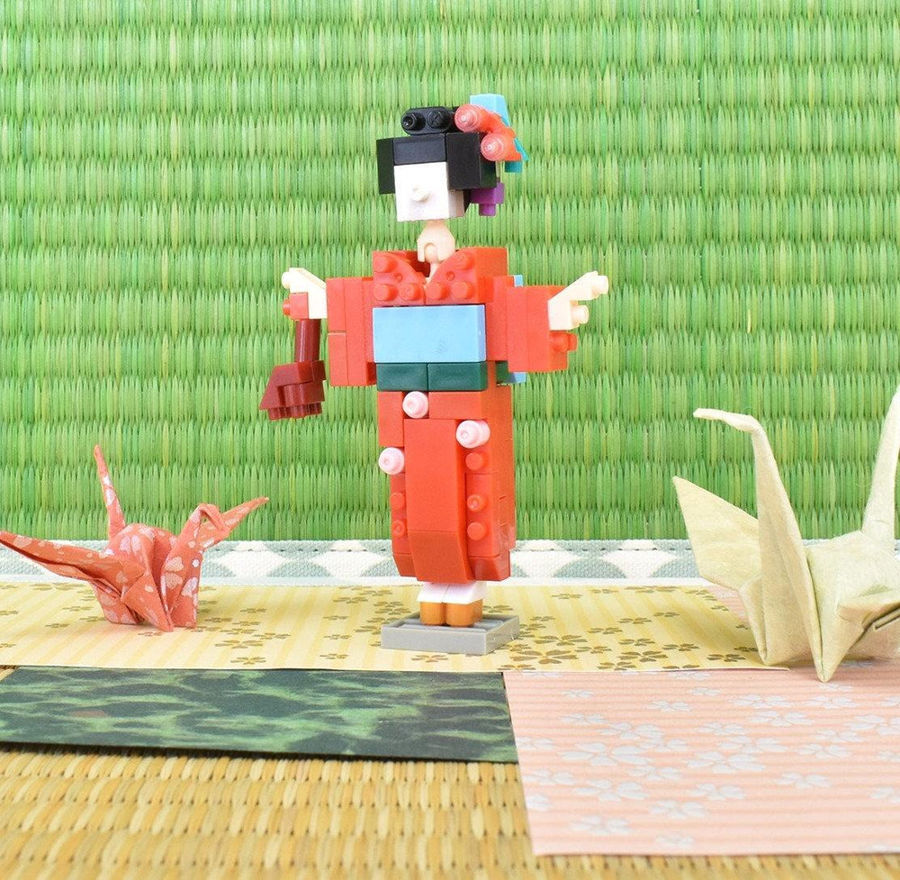 A colorful origami doll dressed in a traditional Japanese kimono stands on a woven mat, surrounded by other origami creations.